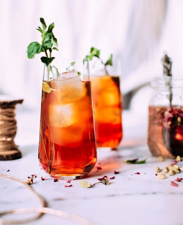 Two tall glass cups filled with a mixed cocktail of bourbon and black tea. It has ice cubes in the glasses and a mint garnish on top of cups.