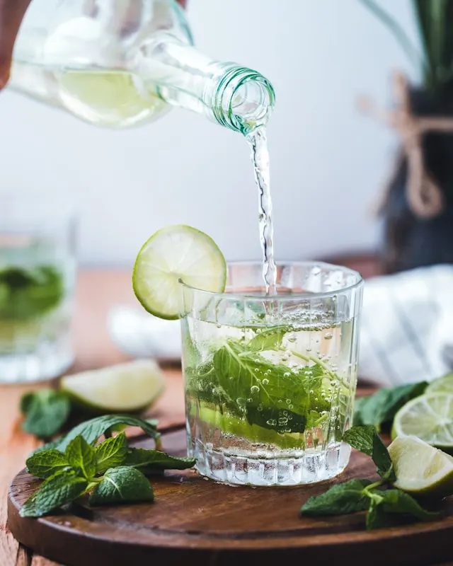 A bottle pouring alcohol in a shot glass with mint leaves and slices of lime inside the cup.