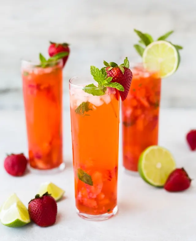 Three tall glass cups filled with strawberry sweet tea, along with multiple red ripe strawberries, lime chunks, ice cubes, and a sprinkle of basil leaves on the cups.