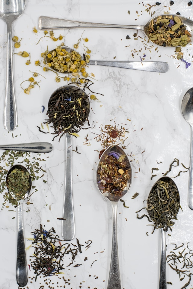 Many different types of loose tea leaves in big silver spoons.